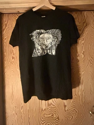 Svart t-shirt med konstnärligt tryck - Svart t-shirt med ett unikt, konstnärligt tryck i vitt på bröstet som föreställer ett abstrakt ansikte med detaljerade linjer och former. Klassisk passform och rund halsringning. Perfekt för dig som vill sticka ut med en cool och kreativ look.