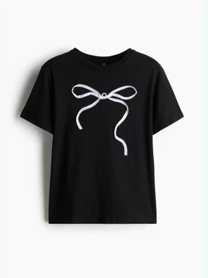 Svart t-shirt med rosettdetalj - Säljer en svart t-shirt i bomull med korta ärmar och en vit rosettdetalj framtill. Enkel och stilren design som passar perfekt till jeans eller kjol. Skön passform och mjukt material.