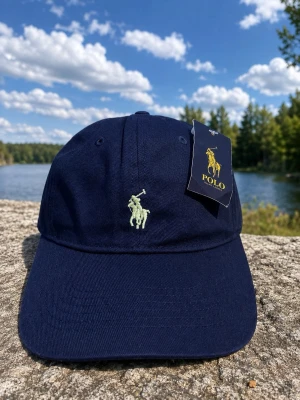 Mörkblå keps från Polo Ralph Lauren - Snygg mörkblå keps från Polo Ralph Lauren med klassisk böjd skärm och broderad logga i ljusgrönt framtill. Kepsen är i bomull och har justerbar rem baktill för perfekt passform. Perfekt accessoar för en sportig och stilren look. Same cap same look the tags inside might be tot eh older model still same cap 