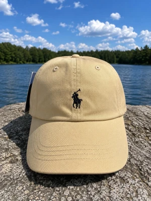 Beige keps från Polo Ralph Lauren - Snygg beige keps från Polo Ralph Lauren med klassisk svart broderad logga framtill och 'POLO' broderat baktill. Justerbar rem bak för perfekt passform. Tillverkad i bomullstyg med böjd skärm och ventilerande öljetter. Tags might be older model still same cap 
