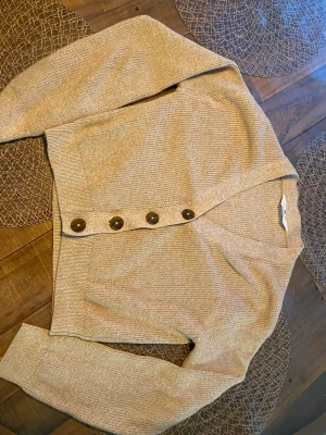 Beige stickad kofta med stora knappar - Snygg beige stickad kofta med fyra stora bruna knappar framtill. Koftan har v-ringning, lång ärm och ribbstickade muddar. Perfekt att slänga över en t-shirt eller linne för en chill look.