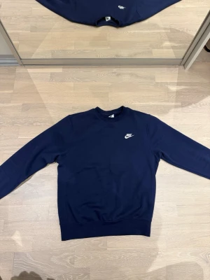 Mörkblå crewneck från Nike - Mörkblå sweatshirt från Nike med klassisk vit logga på bröstet. Tröjan har rund halsringning, långa ärmar och ribbade muddar vid ärmslut och nederkant. Perfekt för en avslappnad och sportig stil.