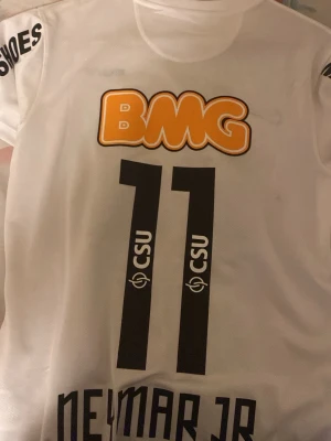 Vit fotbollströja Neymar Jr 11 - Säljer en vit fotbollströja med Neymar Jr och nummer 11 på ryggen. Tröjan har svarta detaljer, tryckta sponsorer som BMG och CSU, samt korta ärmar. Perfekt för dig som gillar fotboll och vill ha en snygg supportertröja.