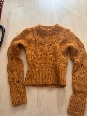 Stickad tröja i ull/alpackablandning XS - Säljer en snygg stickad tröja i varm orange färg, gjord i en mjuk ull- och alpackablandning. Tröjan har hålmönster, ribbade muddar och en rund halsringning. Perfekt för dig som gillar trendiga och mysiga plagg.