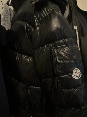 Svart pufferjacka från Moncler - Säljer en svart pufferjacka från Moncler med glansig finish och klassisk Moncler-logga på ärmen. Jackan har huva, dragkedja och fickor framtill. Perfekt för kalla vinterdagar och riktigt snygg streetstil.
