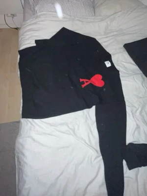 Svart stickad tröja från Ami Paris - Snygg svart stickad tröja från Ami Paris med deras ikoniska röda hjärta och A-logga broderad på bröstet. Tröjan har lång ärm och klassisk passform, perfekt för en stilren look. Materialet är mjukt och bekvämt.