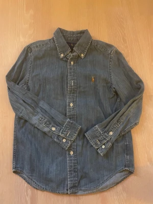 Blå jeansskjorta från Ralph Lauren - Snygg blå jeansskjorta från Ralph Lauren med klassisk krage och knappar framtill. Skjortan har lång ärm och den ikoniska broderade loggan på bröstet. Perfekt till både jeans och chinos för en avslappnad men stilren look.