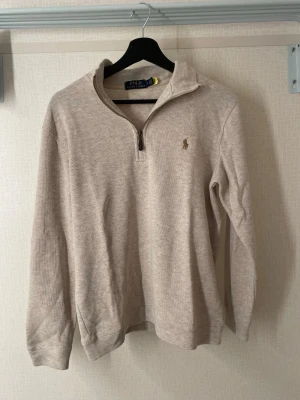 Beige halvzip från Polo Ralph Lauren - Snygg beige tröja från Polo Ralph Lauren med halv dragkedja och klassisk broderad logga på bröstet. Tillverkad i mjukt material och har långa ärmar samt ribbade muddar. Perfekt för en avslappnad men stilren look.