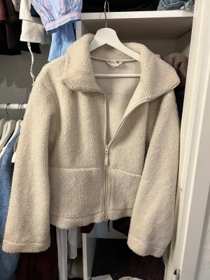 Beige teddyjacka med dragkedja - Mysig beige teddyjacka med hög krage och hel dragkedja framtill. Jackan har två stora fickor och är gjord i fluffigt, mjukt material som ger en avslappnad och trendig look!