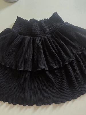 Svart volangkjol från Kappahl - Supersöt svart kjol från Kappahl med bred resår i midjan och dubbla volanger. Kjolen har en plisserad look och är perfekt för dig som gillar en lekfull och trendig stil.