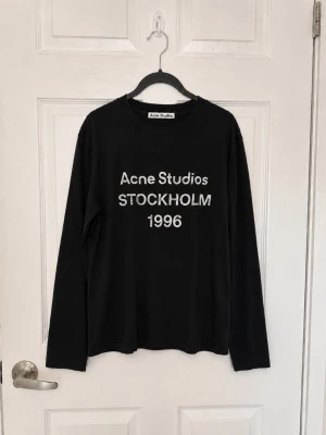 Svart långärmad t-shirt Acne Studios - Svart långärmad t-shirt från Acne Studios med vit text 'Acne Studios STOCKHOLM 1996' på bröstet. Klassisk rund halsringning och rak passform. Perfekt för en stilren och trendig look.