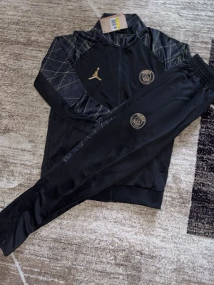 Svart PSG träningsset med guldlogga - Snyggt svart träningsset med jacka och byxor från Paris Saint-Germain. Setet har guldiga detaljer, inklusive Jordan-logga och PSG-emblem på både jacka och byxa. Jackan har mönstrade axelpartier och dragkedja framtill. Perfekt för fotbollsträning eller chill.