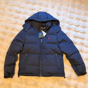 Polo Ralph Lauren Vinterjacka - Snygg mörkblå dunjacka från Polo Ralph Lauren med huva, dragkedja framtill och tre praktiska fickor med dragkedja. Jackan har det klassiska röda Polo-logotypen på bröstet och en puffig, varm design perfekt för kalla vinterdagar.