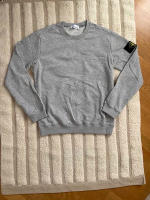 Grå sweatshirt från Stone Island - Säljer en stilren grå sweatshirt från Stone Island med den klassiska loggan på vänster ärm. Tröjan har rund halsringning, ribbade muddar och är tillverkad i mjukt material som passar perfekt till en avslappnad look.