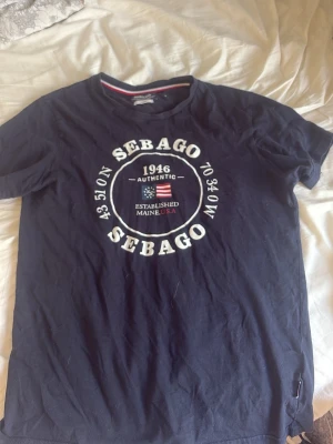 Mörkblå Sebago t-shirt med tryck - Snygg mörkblå t-shirt från Sebago med stort vitt tryck framtill och detaljer i rött. Klassisk rund halsringning och korta ärmar. Tillverkad i mjuk bomull som känns skön mot huden. Perfekt för en avslappnad och stilren look.