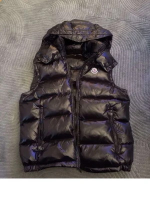 Svart dunväst från Moncler med luva - Snygg svart dunväst från Moncler med glansig finish och avtagbar luva. Västen har två dragkedjeförsedda fickor framtill och klassisk Moncler-logga på bröstet. Insidan har ett coolt serietryck och Moncler-märke. Perfekt för lager på lager och streetstyle.