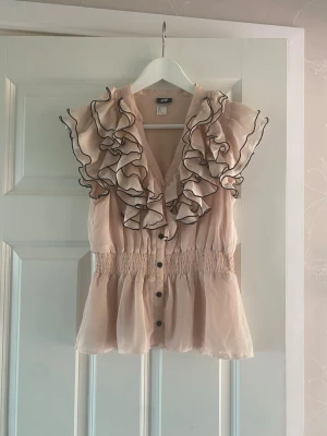 Rosa volangblus från H&M - Supersöt beige blus från H&M med volanger och svarta kanter längs volangerna. Blusen har knappar framtill, smock i midjan och är tillverkad i ett tunt, lätt material som ger ett luftigt intryck.
