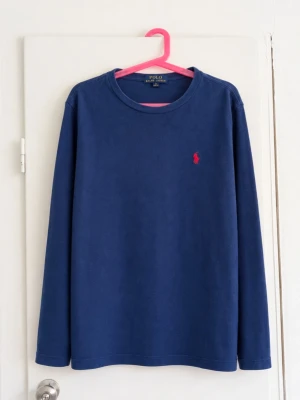 Polo Ralph Lauren långärmad tröja i klassiskt mörkblått 💙 - Stilren modell med rund hals och den klassiska röda broderade Polo-loggan på bröstet. Enkel och lättmatchad – perfekt till jeans, chinos eller under en hoodie/jacka. • Märke: Polo Ralph Lauren • Storlek: M (10–12 år) • Färg: Mörkblå • Långärmad • Rund hals • Röd broderad Polo-logga framtill Fint begagnat skick utan defekter!