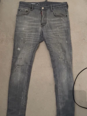  Dsquared2 jeans  - Säljer ett par grå jeans från Dsquared2 i modell med smal passform. Jeansen har slitna detaljer och diskreta färgstänk för en cool, trendig look. Klassisk femficksmodell med knappgylf och logotypdetalj vid fickan. Äkta 