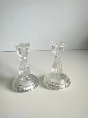 Två ljusstakar i räfflat glas - Två stilrena ljusstakar i transparent pressglas med dekorativa räfflade mönster och rund fot. Klassisk design som passar i de flesta rum. 10cm höga. Mycket gott skick!