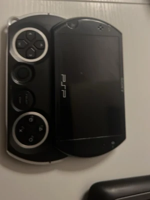 Sony PSP Go bärbar spelkonsol svart - Säljer en svart Sony PSP Go, en smidig och kompakt bärbar spelkonsol med utfällbar skärm och klassiska PlayStation-knappar. Konsolen har en stilren design i plast och metall, perfekt för gaming på språng.