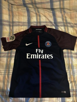 PSG matchtröja Nike mörkblå - Snygg mörkblå Paris Saint-Germain matchtröja från Nike med röda detaljer och klubbmärke på bröstet. Tröjan har korta ärmar i vinrött och vit Fly Emirates-logga på magen. Perfekt för fotbollsfans som vill representera PSG.