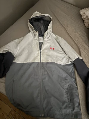 Gråvit vindjacka från Under Armour - Snygg vindjacka från Under Armour i tvåfärgad design med vit ovandel och grå nederdel. Jackan har huva, dragkedja framtill, två sidofickor och svart Under Armour-logga på bröstet. Perfekt för blåsiga dagar och sportiga outfits.