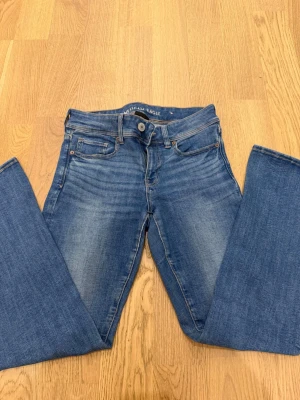 Blå bootcut jeans American Eagle - Snygga blå bootcut jeans från American Eagle med klassisk femficksdesign och lätt slitna detaljer. Jeansen har normalhög midja och är tillverkade i stretchigt denim för en bekväm passform. Perfekta för en avslappnad och trendig look. Det är short. 