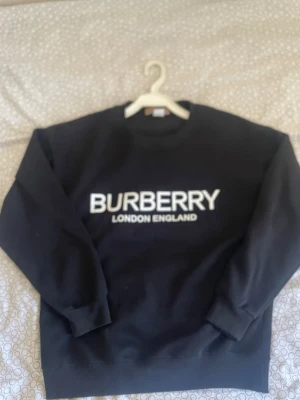 Svart sweatshirt från Burberry - Snygg svart sweatshirt från Burberry med stor vit logga och texten 'London England' på bröstet. Klassisk rund halsringning och långa ärmar. Perfekt för dig som gillar stilrena och exklusiva plagg.