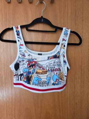 Vit crop top med serietidningsmönster - Cool vit croppad linnetopp med färgglatt serietidningsinspirerat tryck och röda detaljer längst ner. Toppen har breda axelband och ärmlös design, perfekt för en avslappnad och trendig look.