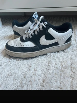 Svarta och vita Nike sneakers - Säljer ett par klassiska Nike sneakers i svart och vitt läder med vit sula och snörning. Skorna har den ikoniska swoosh-loggan på sidan och NIKE-text bak på hälen. Perfekta för dig som gillar stilrena och tidlösa sneakers.
