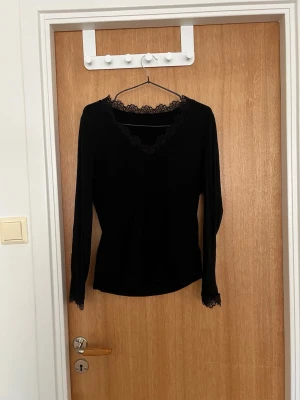 Svart långärmad topp med spets - Snygg svart långärmad topp med v-ringning och fina spetsdetaljer vid halsringningen och ärmsluten. Perfekt att styla med jeans eller kjol för en trendig look.
