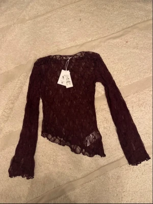 Vinröd spetsblus med asymmetrisk kant - Vinröd långärmad blus i spets med asymmetrisk nederkant och transparent design. Blusen har smal passform och fina volangdetaljer vid ärmslut och nederkant. Perfekt för dig som vill ha en unik och trendig topp.
