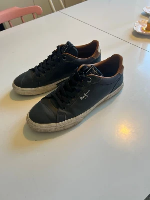Mörkblå sneakers från Pepe Jeans - Mörkblå sneakers från Pepe Jeans med bruna detaljer vid hälen och vita sulor. Skorna har klassisk låg profil, svarta skosnören och diskret logga på sidan. Materialet är slätt läderliknande och insidan har en kontrasterande brun färg.