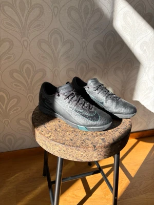 Svarta Nike futsasl/ inomhus skor - Snygga svarta fotbollsskor med turkosa detaljer och mönstrad ovandel. Skorna har klassisk snörning och låg profil, perfekt för snabba rörelser på planen. Tillverkade i syntetmaterial för bra grepp och kontroll.