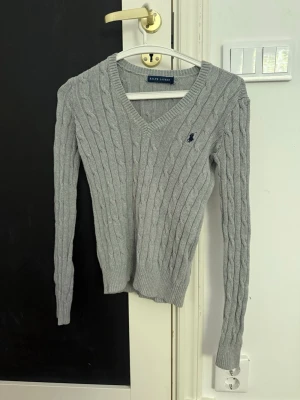 Grå kabelstickad tröja Ralph Lauren - Kabelstickad grå tröja från Ralph Lauren med V-ringning och lång ärm. Tröjan har en liten mörkblå logga broderad på bröstet och ribbade muddar vid ärmslut och nederkant. Klassisk och tidlös design i mjukt material.