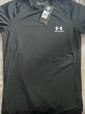 Svart träningströja Under Armour - Snygg svart träningströja från Under Armour med logga och text i vitt på bröstet. Tillverkad i ett stretchigt funktionsmaterial som andas, perfekt för gym eller löpning. Kortärmad modell med rund hals och smal passform.