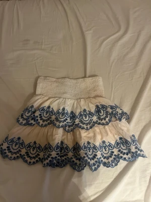 Vit kjol med blå broderade volanger - Söt kortkjol i vitt med två lager volanger och blå broderade detaljer. Kjolen har bred resår i midjan för bekväm passform och är perfekt för sommarens alla tillfällen. Materialet känns lätt och luftigt.