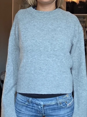 gina Ljusgrå stickad tröja - Mysig ljusgrå stickad tröja med rund halsringning och ribbade muddar vid ärmslut och nederkant. Tröjan har en rak och något boxig passform, perfekt att styla med jeans eller kjol. Materialet ser mjukt och bekvämt ut.