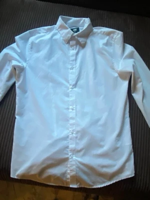 Vit slim fit skjorta från H&M - Klassisk vit skjorta från H&M i slim fit-modell. Skjortan har långa ärmar, knappar framtill och en stilren krage. Perfekt för dig som gillar en clean och enkel look. Materialet är bomull och den är lätt att stryka.