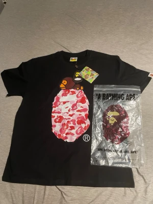 Svart BAPE t-shirt med camotryck - Säljer en svart t-shirt från A Bathing Ape med ikoniskt BAPE-tryck framtill i rosa, rött och vitt camomönster samt apmotiv. Klassisk rund hals och korta ärmar. Perfekt för dig som gillar streetwear och vill sticka ut med en unik design.