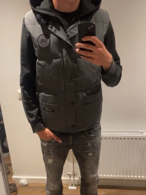 Grå dunväst från Canada Goose - Snygg grå dunväst från Canada Goose med två stora fickor framtill. Västen har diskret logga på bröstet och stängs med knappar. Perfekt för lager-på-lager och håller dig varm under kyliga dagar. Använd 2 gånger. 