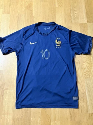 Blå Frankrike fotbollströja Nike vm 22 - Säljer en blå officiell Frankrike fotbollströja från Nike med nummer 10 och Mbappé på ryggen. Tröjan har korta ärmar, guldiga detaljer och FFF-logga med tupp och två stjärnor på bröstet. Perfekt för dig som älskar fotboll och vill stötta landslaget. Storleken är L men passar både L och M