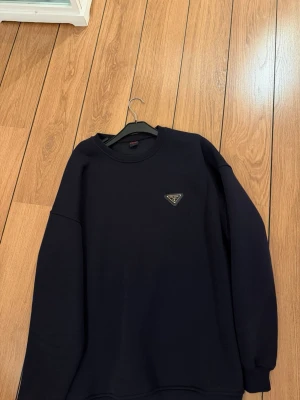 Mörkblå sweatshirt från Prada - Snygg mörkblå sweatshirt från Prada med klassisk rund halsringning och diskret logotyp på bröstet. Tröjan har ribbade muddar vid ärmslut och nederkant. Tillverkad i mjukt material som känns skönt mot huden. Perfekt för en clean och stilren look.