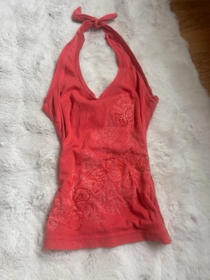 Korallfärgad halterneck - Snygg korallfärgad halterneck topp med knyt i nacken. Toppen har blommigt mönster och detaljer med pärlor framtill. Perfekt för sommarens alla tillfällen och ger en fräsch look till din outfit. Sydd till halterneck själv, pris går att diskuteras💕