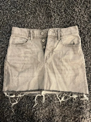 Grå jeanskjol med fransig kant - Säljer en grå jeanskjol i kort modell med rå, fransig kant nedtill. Kjolen har knappgylf, bälteshällor och klassiska fem fickor. Perfekt för dig som gillar en avslappnad och trendig stil.