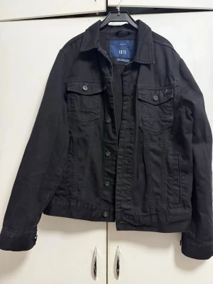 Svart jeansjacka från 1975 - Stilren svart jeansjacka från 1975 med klassisk krage, två bröstfickor med lock och knappar samt sidofickor. Jackan har knappstängning framtill och justerbara ärmslut. Perfekt för dig som gillar en enkel men cool look.