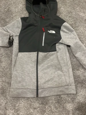 Grå hoodie från The North Face - Snygg grå hoodie från The North Face med svart panel upptill och röd dragkedja. Har huva, hel dragkedja framtill och en bröstficka med dragkedja. Perfekt för dig som gillar sportig och avslappnad stil.