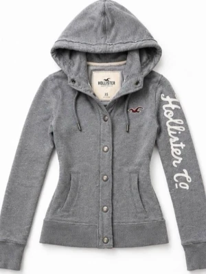 Grå hoodie från Hollister - Snygg grå hoodie från Hollister med knappstängning framtill, dragsko i huvan och Hollister Co.-logga i vitt längs ena ärmen. Klassisk Hollister-fågel broderad på bröstet. Mjuk bomullsmix och ribbade muddar vid ärmslut och nederkant.