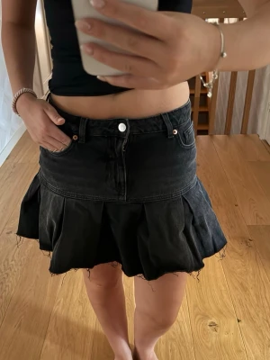 Svart jeanskjol med volang - Säljer en svart kort jeanskjol med volangkant och råa kanter. Kjolen har klassiska jeansdetaljer som fickor och knappstängning framtill. Perfekt för dig som gillar en edgy och trendig stil.